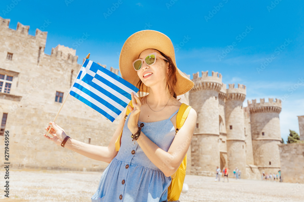 Happy asian traveler woman with greek flag enjoying great view to the old Knights Grand Master Castle in Rhodes, Greece