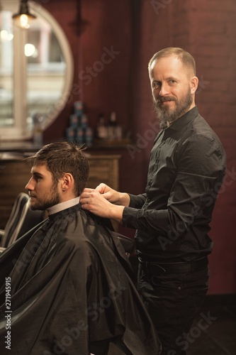 Smiling male barber looking at camera while preparing client for new hairstyle in vintage barbershop. Man wearing black gown sitting in chair and looking at mirror. Concept of service and haircut.