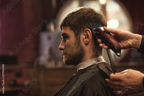 Side view of male client in barbershop wearing gown sitting in chair and looking at mirror. Hand of male hairdresser keeping trimmer and doing stylish haircut. Concept of hairstyle.