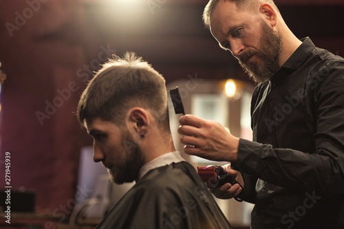 View from side of serious barber doing new haircut to male client sitting in chair in barbershop.Professional hairdresser keeping trimmer and brushing hair with comb. Concept of trendy hairstyle.