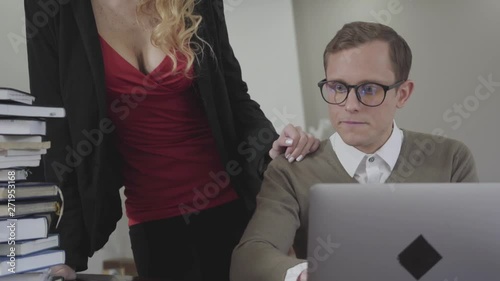 Young modestly dressed man sitting at table working on laptop. Hot woman with deep neckline bends over and puts hand on his shoulder. The nerd timidly takes her hand off. Secretary seduces boss
