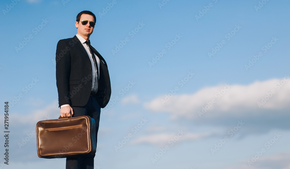 Rich, stylish businessman in glasses, in a suit and with a suitcase stands against the blue sky. Business, work and a lot of money. Oligarch, director, chief. Waiting for transport. Rich, stylish businessman in glasses, in a suit and with a suitcase stands against the blue sky. Business, work and a lot of money. Oligarch, director, chief. Waiting for transport.
