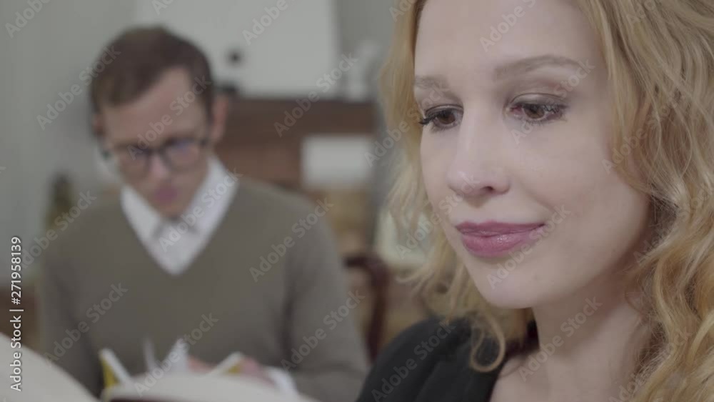 Close-up face of beautiful blond woman reading aloud the book in the foreground while modestly dressed man looking at the lady on the background. The nerd and hot woman working together Close-up face of beautiful blond woman reading aloud the book in the foreground while modestly dressed man looking at the lady on the background. The nerd and hot woman working together
