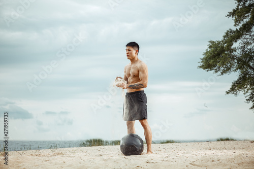 Young healthy man athlete doing exercise with ball at the beach. Signle male model shirtless training air at the river side in sunny day. Concept of healthy lifestyle, sport, fitness, bodybuilding. photo