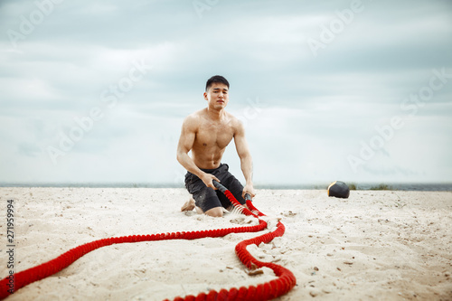 Young healthy man athlete doing exercise with the rope at the beach. Signle male model shirtless training air at the river side in sunny day. Concept of healthy lifestyle, sport, fitness, bodybuilding photo