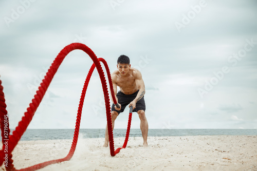 Young healthy man athlete doing exercise with the rope at the beach. Signle male model shirtless training air at the river side in sunny day. Concept of healthy lifestyle, sport, fitness, bodybuilding photo