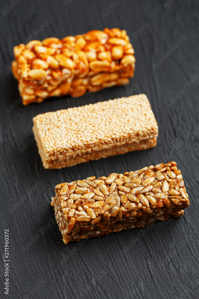 Cereal granola bar with peanuts, sesame and sunflower seeds on a cutting board on a dark stone table. View from above. Three Assorted Bars Cereal granola bar with peanuts, sesame and sunflower seeds on a cutting board on a dark stone table. View from above. Three Assorted Bars