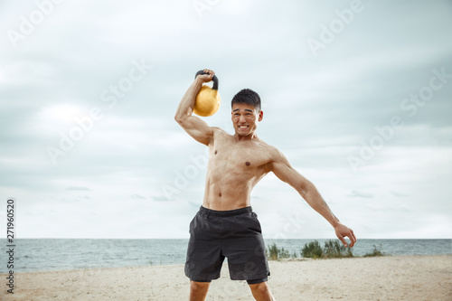 Young healthy man athlete doing exercise with the weight at the beach. Signle male model shirtless training at the river side in sunny day. Concept of healthy lifestyle, sport, fitness, bodybuilding. photo