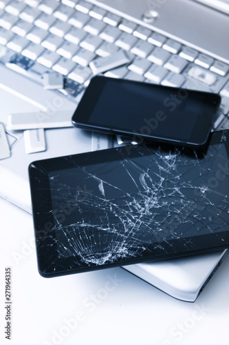 A silver laptop with a broken keyboard, tablet with a cracked display and black phone. A close-up picture of part of broken laptop and cracked screen on a tablet.