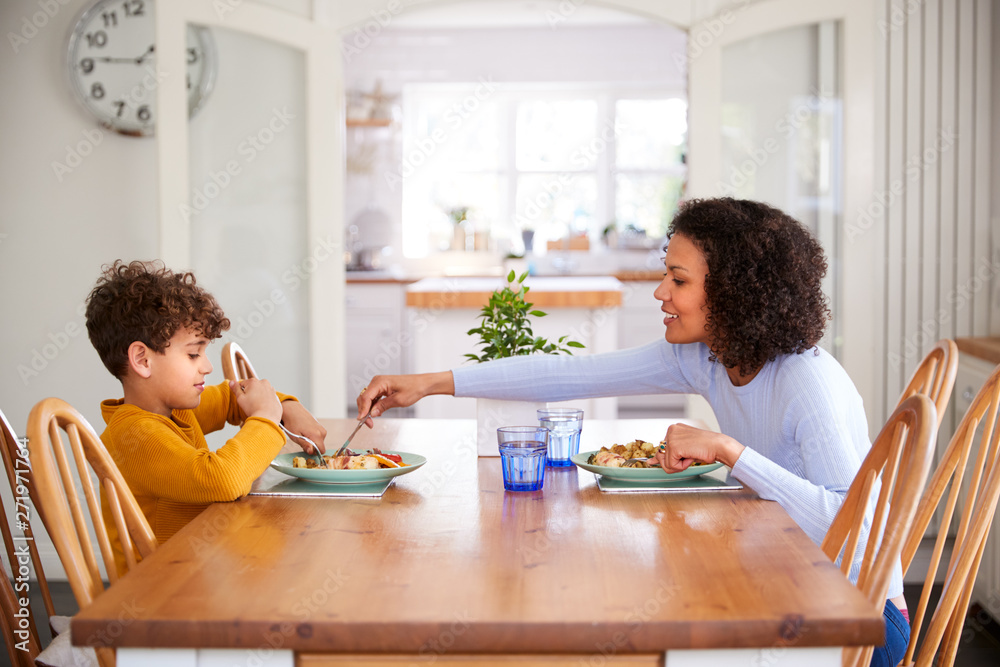 Single Mother Sitting At Table Eating Meal With Son In Kitchen At Home Single Mother Sitting At Table Eating Meal With Son In Kitchen At Home