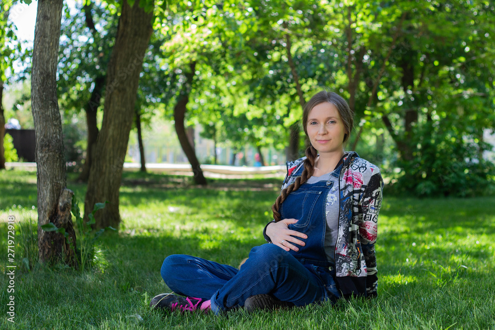 beautiful woman in early pregnancy in denim jumpsuit sitting on the green grass in the Park beautiful woman in early pregnancy in denim jumpsuit sitting on the green grass in the Park