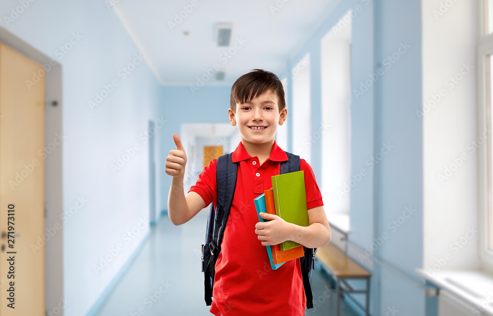 education and people concept - smiling little student boy in red polo t-shirt in glasses with books and bag showing thumbs up over school corridor background education and people concept - smiling little student boy in red polo t-shirt in glasses with books and bag showing thumbs up over school corridor background