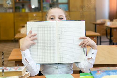 The schoolgirl covered her face with a school gradebook. A teenager girl hides her face behind an open book raised by a school book. photo