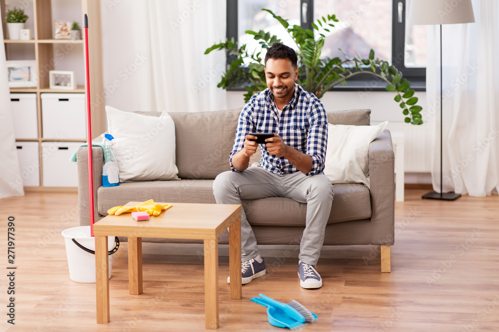 household and technology concept - indian man playing game on smartphone after cleaning home