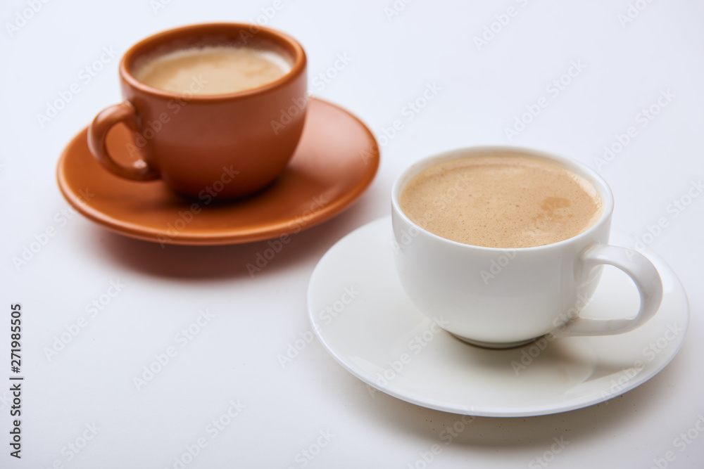 selective focus of delicious coffee with foam in cups on saucers on white background selective focus of delicious coffee with foam in cups on saucers on white background