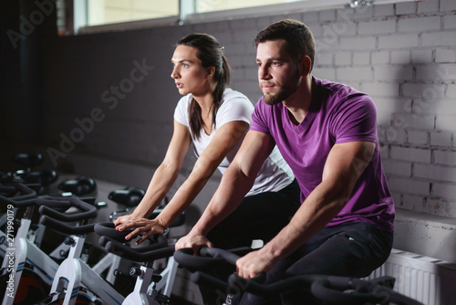 Attractive woman and man biking in the gym, exercising legs doing cardio workout cycling bikes. Couple in a spinning class wearing sportswear. Fitness healthy lifestyle concept © ANR Production