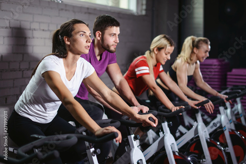Group of smiling friends at gym exercising on stationary bike. Happy cheerful athletes training on exercise bike. Young men and woman working out at a class in the gym.