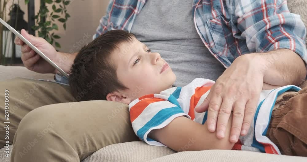 Father reading ebook story to his son trying to sleep on his lap at home