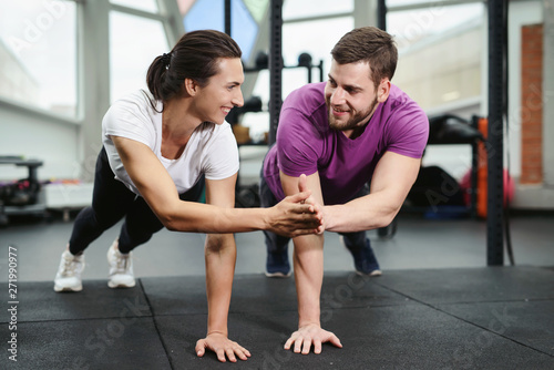 Young athletic man and woman are doing plank together and holding hand to hand in the gym. Sporty couple planking and giving high five. Sport fitness exercises in health club
