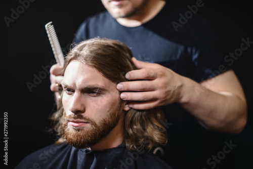 Handsome bearded man, hipster with beard and moustache has haircut in hairdressing saloon or barbershop. Barber works with hair trimmer or clipper. Black background isolated