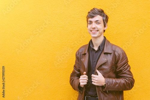 Photos Young smiling handsome guy with brown greyish hair in a brown leather jacket and black shirt staying against the bright yellow wall
