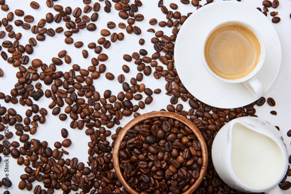 Obraz premium top view of tasty coffee in cup on saucer near scattered roasted beans, wooden bowl and milk jug