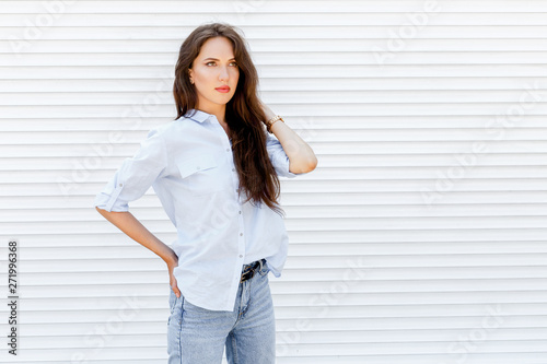 Young stylish beautiful brunette woman wearing blue shirt and denim jeans posing against white street wall. Trendy spring summer casual outfit. Street fashion.