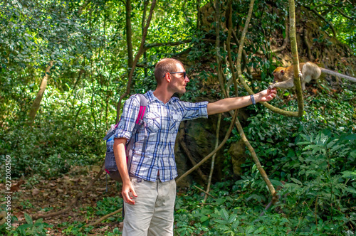Young man with backpack reaching his hand to macaque. European traveler is feeding monkey in jungle. Baby monkey sitting on tree and catching banana.