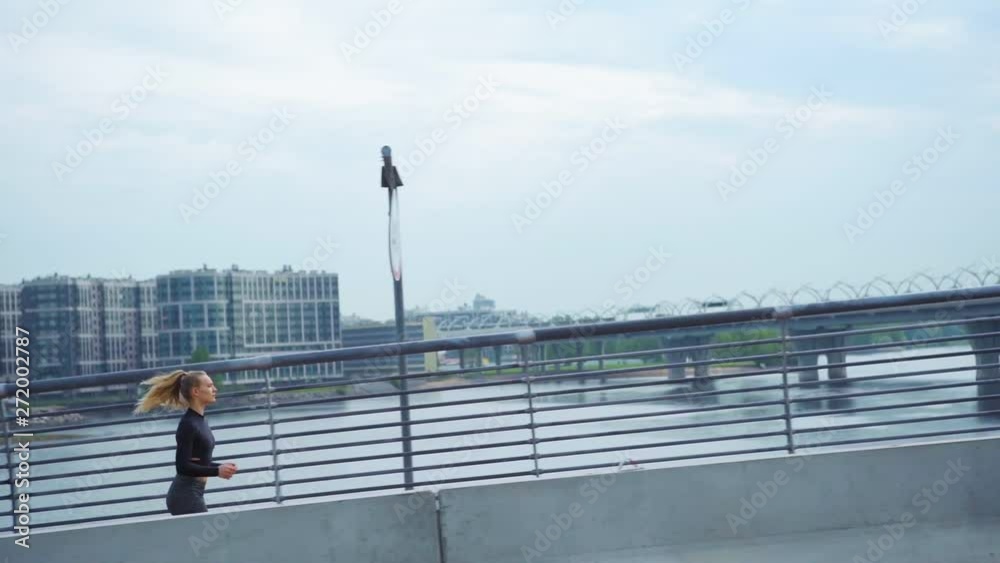 Tracking right of determined female jogger running up on bridge road over water during marathon training, side view slow motion shot