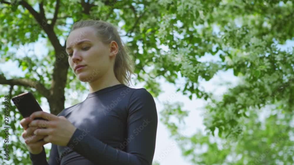 Low angle view of worried female athlete in cropped top standing under blooming trees in park and texting on cell phone after training, panning slow motion shot Low angle view of worried female athlete in cropped top standing under blooming trees in park and texting on cell phone after training, panning slow motion shot