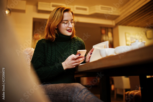 Cheerful young beautiful redhaired woman in glasses, green warm wool sweater, using her phone and notebook, while sitting at her working place on cafe.