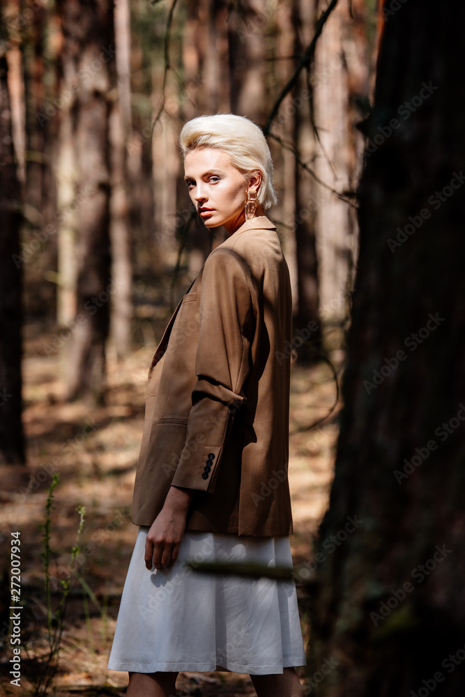 elegant serious blonde woman looking at camera while standing in forest