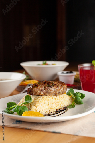 Close up of a roasted pork cutlet with boiled bulgur and greens as a part of set meal in a restaurant, with other meals on a background