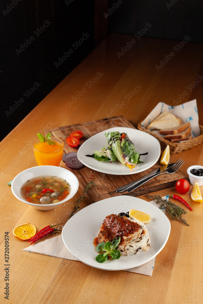Lunching out in a restaurant: fish with steamed rice, light vegetable soup with meatballs and vegetarian spring roll on a dark background Lunching out in a restaurant: fish with steamed rice, light vegetable soup with meatballs and vegetarian spring roll on a dark background