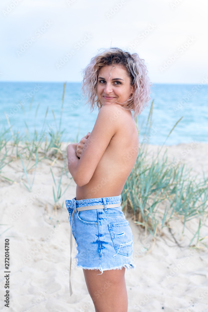 beautiful young woman with curly hairs in denim shorts and swimsuit is happy to be on the beach near the ocean. Ocean and grass on the background beautiful young woman with curly hairs in denim shorts and swimsuit is happy to be on the beach near the ocean. Ocean and grass on the background