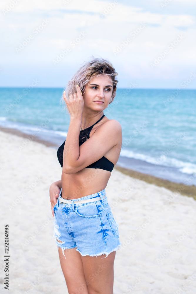 beautiful young woman with curly hairs in denim shorts and swimsuit is happy to be on the beach near the ocean. Ocean and grass on the background beautiful young woman with curly hairs in denim shorts and swimsuit is happy to be on the beach near the ocean. Ocean and grass on the background
