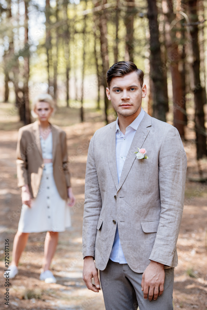 stylish groom with boutonniere on jacket and bridegroom standing in forest and looking at camera stylish groom with boutonniere on jacket and bridegroom standing in forest and looking at camera