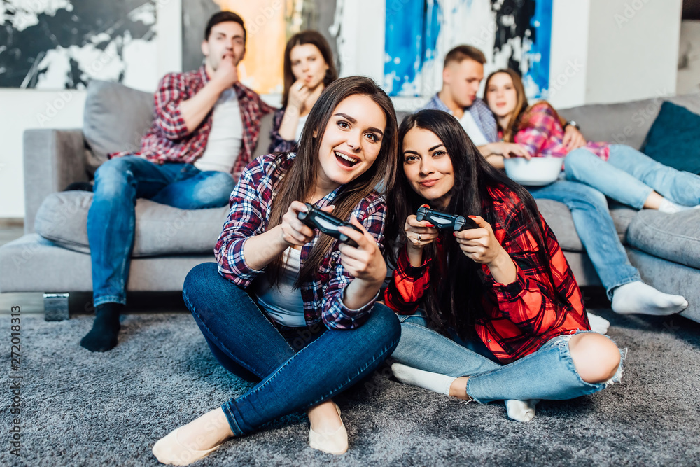 Obraz premium Two young, brunette woman playing video games at home with their company behind..