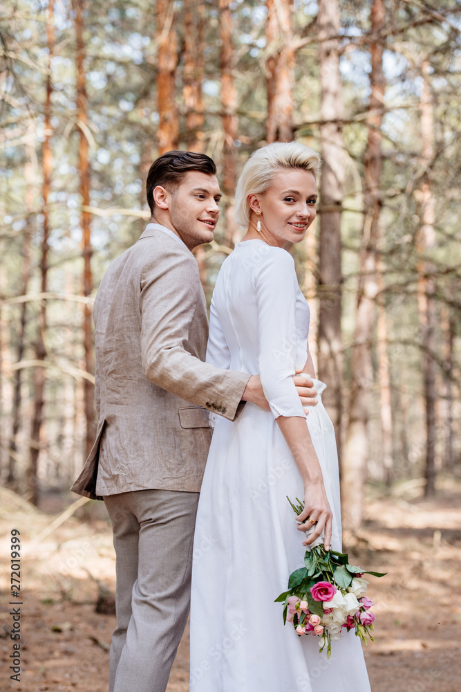 smiling bridegroom in formal wear embracing bride in forest