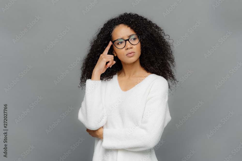 Φωτογραφία Troubled serious female keeps fore finger on temple, looks aside, has curly hair, being deep in thoughts, wears white jumper and optical glasses, isolated on grey background