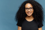Headshot of pleasant looking dark skinned woman with Afro haircut, wears transparent glasses and casual black t shirt, expresses good emotions, stands against blue background, free space for slogan