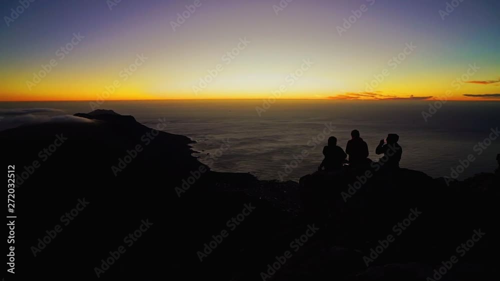 A silhouette of 3 young friends drinking sundowners on top of a mountain as the clouds roll in beneath them with an orange setting sun. Table Mountain, Cape Town, South Africa. A silhouette of 3 young friends drinking sundowners on top of a mountain as the clouds roll in beneath them with an orange setting sun. Table Mountain, Cape Town, South Africa.