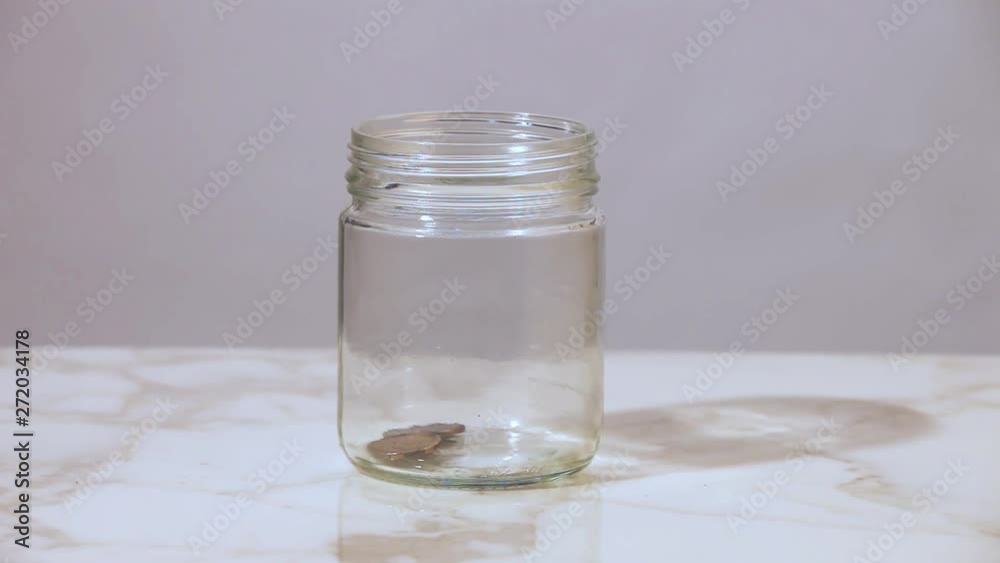 Coins being dropped intro a glass jar on a white table. Coins being dropped intro a glass jar on a white table.