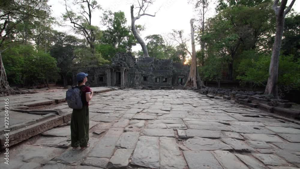 Caucasian woman with backpack is looking at Ta Prohm temple in the evening. Angkor Wat Complex, Cambodia Caucasian woman with backpack is looking at Ta Prohm temple in the evening. Angkor Wat Complex, Cambodia
