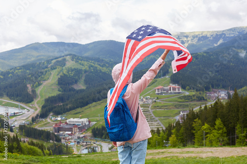 Experienced traveller wearing jeans, pink jacket with hood and blue backpack, raising big U. S. flag up, feeling of freedom, travel tour, go camping in mountains. Free time activities concept. photo