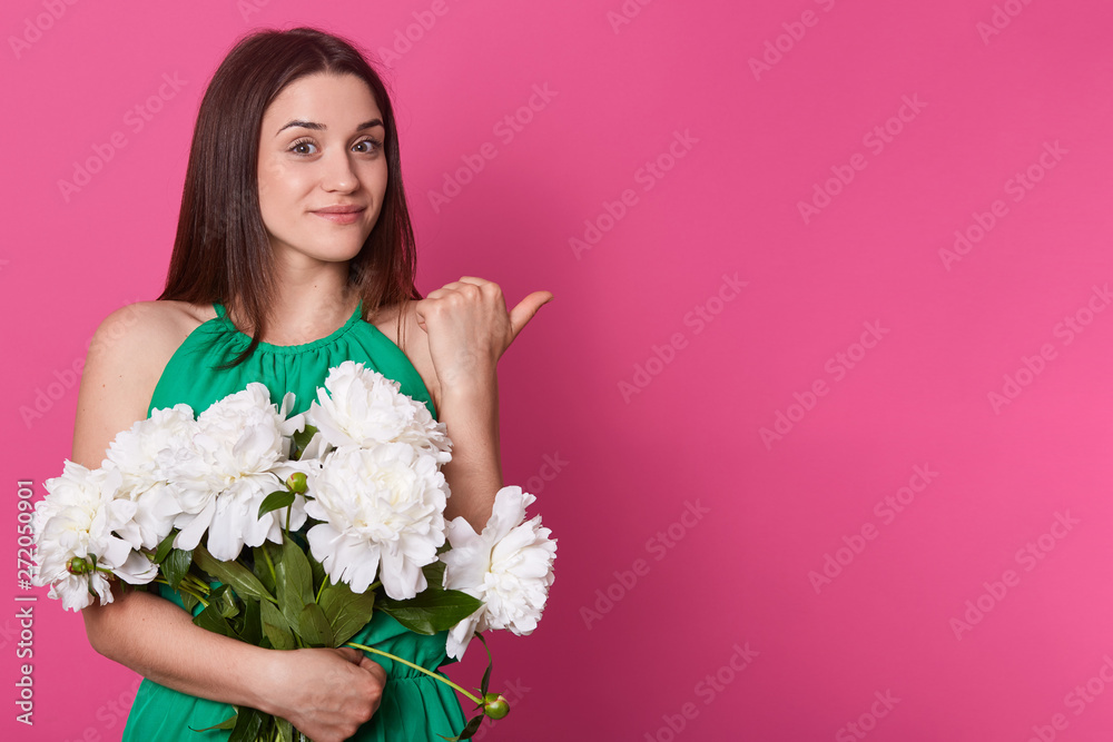 Beautiful pleasant young female looking directly at camera, smiling sweet, making gesture with her thumb aback, holding big bouquet of white peonies, wearing green dress. Copyspace for advertisement.