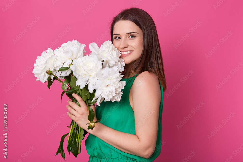 Portrait of cheerful tender model standing isolated over pink background in studio, smelling and putting close to face peonies, being in high spirits, getting present. People and emotions concept. Portrait of cheerful tender model standing isolated over pink background in studio, smelling and putting close to face peonies, being in high spirits, getting present. People and emotions concept.