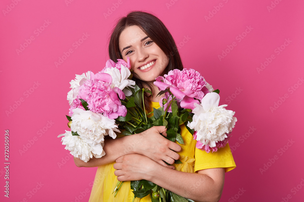 Portrait of positive energetic female holding spring flowers close to her body, looking directly at camera, having sincere smile, flower lover, getting pleasant gifts, wearing bright yellow dress.