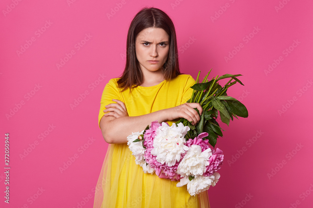Indoor studio shot of charismatic model with frowned face and folded arms, holding bouquet of white and pink flowers, going to throw them out, wearing yellow dress, isolated over pink background. Indoor studio shot of charismatic model with frowned face and folded arms, holding bouquet of white and pink flowers, going to throw them out, wearing yellow dress, isolated over pink background.