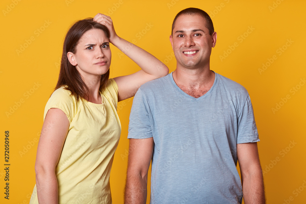 Close up portrait of couple in love, dissatisfied woman keeps hand on male's shoulder, handsome man stands smiling, looking directly at camera, yuoung family posing isolated over yellow background. Close up portrait of couple in love, dissatisfied woman keeps hand on male's shoulder, handsome man stands smiling, looking directly at camera, yuoung family posing isolated over yellow background.
